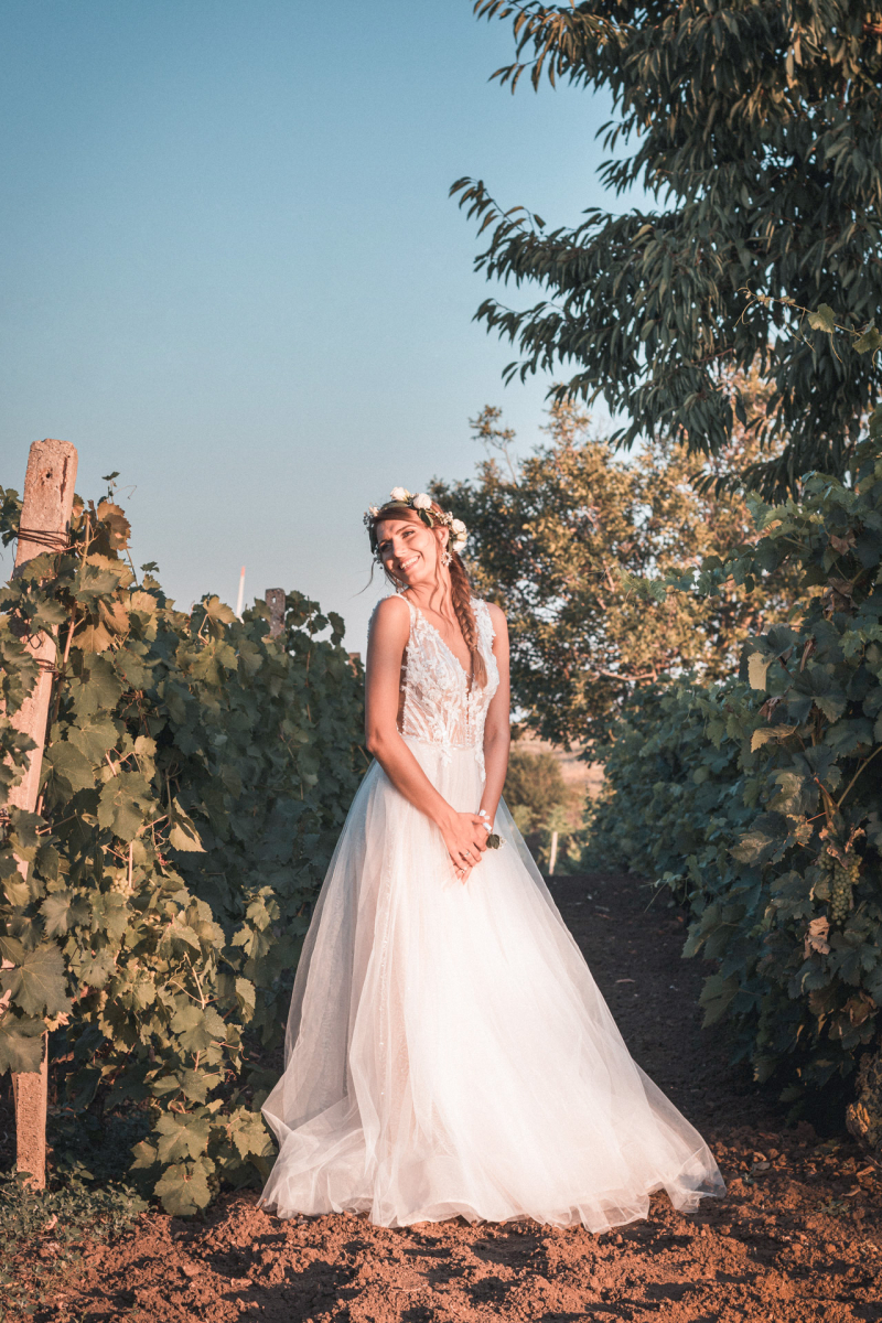 Усмихната булка в дантелена рокля и венец, позираща в лозе при залез | Smiling bride in a lace dress and flower wreath posing in a vineyard at sunset